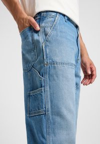 Jeans in denim azzurro chiaro con un fit rilassato, dotati di più tasche, cuciture a vista e dettagli di texture sottili. Mano posizionata in una tasca.