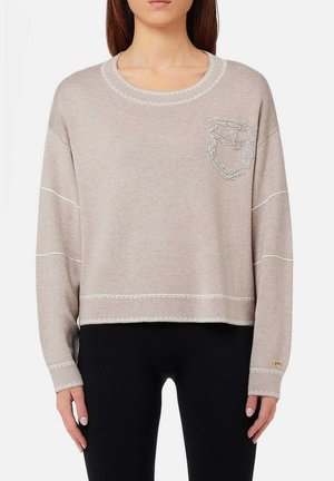 Donna che indossa un maglione beige a maniche lunghe con dettagli di cucitura bianchi e un piccolo taschino texturizzato sul lato sinistro del petto, abbinato a pantaloni neri.