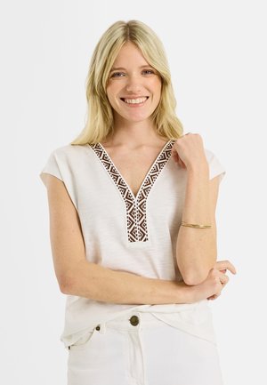 Femme souriante aux longs cheveux blonds portant un haut blanc à col en V marron à motif et un pantalon blanc, posant avec le bras plié et un bracelet en or.