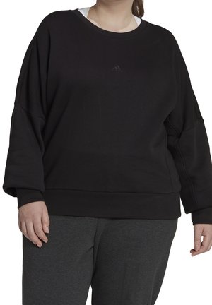 Osoba nosząca czarną bluzę Adidas z małym logo na piersi oraz ciemnoszare spodnie sportowe na białym tle.