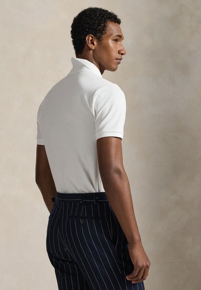 Tânăr cu păr scurt și creț, purtând o tricou polo alb cu mânecă scurtă și pantaloni în dungi bleumarin, privindu-se din profil.