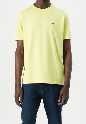 Man draagt lichtgele korte mouw T-shirt met "BOSS" logo op de borst en donkerblauwe spijkerbroek, staand tegen een witte achtergrond.