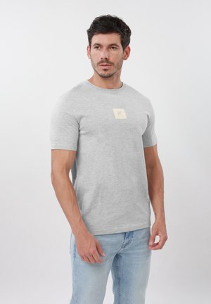 Mand med kort mørkt hår iført en lysegrå T-shirt med en lille beige firkantet logo og lyseblå jeans, stående foran en hvid baggrund.