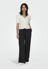 Blouse crème à boutons avec des manches bouffantes, associée à un pantalon noir taille large et des chaussures blanches. Le tissu semble léger et texturé.
