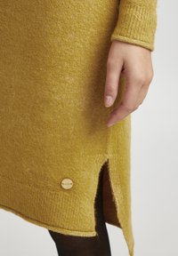 Robe tricotée jaune moutarde avec une finition texturée, fente sur le côté et accent de bouton en bois à l'ourlet. Montre une main reposant sur le tissu.