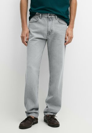 Uomo che indossa jeans grigio chiaro a gamba dritta, scarpe stringate marrone scuro e una camicia verde scuro, in piedi davanti a uno sfondo bianco.