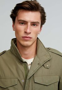 Olive green, waterproof jacket with a high collar, front zip, and button accents. Features two chest pockets and a textured fabric.