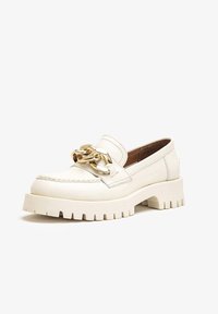 Mocassin blanc épais avec semelle crantée épaisse et grand détail chaîne dorée sur le dessus, incliné vers la gauche, sur fond blanc uni.