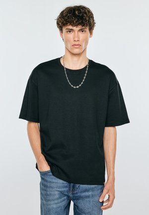 Młody mężczyzna z kręconymi włosami, ubrany w czarny oversize t-shirt, srebrny naszyjnik łańcuszek i niebieskie dżinsy, stoi z jedną ręką w kieszeni.