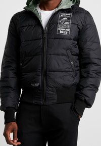 Uomo che indossa una giacca nera imbottita con zip frontale, tasche laterali, polsini a coste e toppe di testo bianco sul petto e sul lato inferiore.