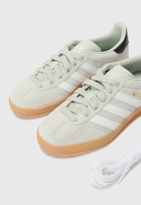 Light grey suede sneakers with white stripes and gold logo. Features a gum rubber sole and includes extra white laces. Textured heel detail.