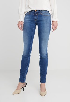 Jeans ajustados de mezclilla azul con un acabado desgastado, cintura de altura media y diseño de largo hasta el tobillo. Combinados con zapatos de tacón alto en color nude.