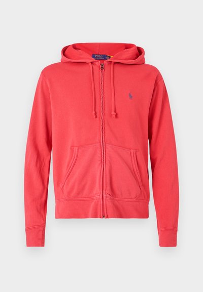 Sudadera roja con cremallera hecha de tela suave, con capucha ajustable, bolsillos frontales y un pequeño logo en el pecho. Textura suave y diseño casual.