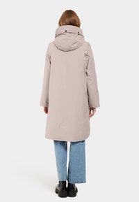 Lungo impermeabile beige con cappuccio, chiusure a scatto e logo, abbinato a jeans chiari a gamba larga e stivali neri chunky.