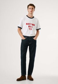 Biela tričko s červeným potlačou "NOTTING HILL 73", námornícke manžety; tmavé džínsy; hnedé semišové chelsea čižmy. Jednoduché pozadie.