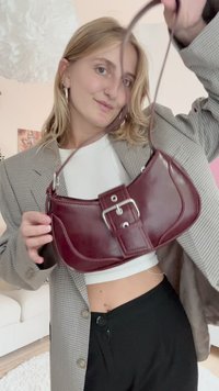 Geantă de mână din piele vișinie cu o formă curbată, accesorii din argint și un accent asemănător unei centuri. Curea ajustabilă, textură neted�ă și design minimalist.