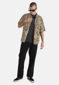 Short-sleeve, light brown floral patterned shirt over a dark t-shirt. Black loose-fit pants and black slip-on shoes. Accessories include sunglasses and rings.