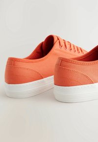 Baskets en toile orange avec un design bas, des semelles en caoutchouc blanches lisses et des œillets assortis pour les lacets. Tissu texturé avec détails de couture.