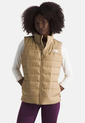 Frau mit lockigem Haar, die eine beige gesteppte Weste über einem weißen Langarmshirt und lila Hosen trägt, mit den Händen in den Westentaschen.