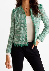 Vihreä tweed-takki hapsureunuksilla, pyöreällä kaula-aukolla ja ilman nappeja. Päällä istuva valkoinen toppi ja tummat housut.