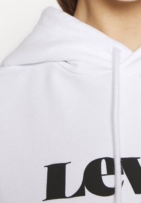 Felpa bianca realizzata in un tessuto morbido, con un grande logo nero "Levi's" stampato sul davanti. Presenta un cappuccio con cordino e una superficie texturizzata.