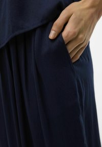 Tamno plavi odjevni predmet glatke, lagane teksture. Odlikuje se slobodnim krojem i bočnom džepom, s širokom siluetom koja se elegantno obavija.