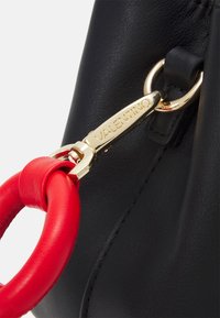 Detail černé kožené kabelky se zlatým popruhem a červeným koženým očkem. Na kování je zobrazen název značky "Valentino."