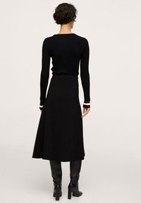 Pull noir côtelé à manches longues, associé à une jupe fluide noire, porté avec des bottes noires montantes. Le pull présente des détails rayés aux poignets.