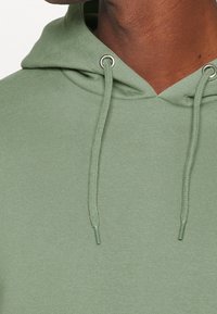 Sweat à capuche vert en tissu doux, doté d'une poche kangourou, de cordons de serrage et d'œillets argentés. Texture lisse avec une coupe décontractée.