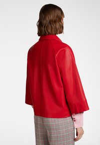 Giacca di pelle rossa con una vestibilità leggermente ampia, maniche larghe e colletto a punte. Abbinata a pantaloni a quadri grigi con motivo.