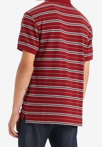 Polo rouge rayé avec des lignes horizontales blanches et noires, manches courtes, col côtelé et fentes sur les côtés à l'ourlet pour faciliter le mouvement.