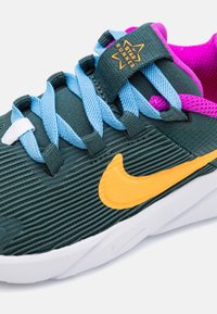 Baskets en tissu vert foncé avec des lacets bleus, un swoosh Nike orange et des accents roses. Surface texturée avec un design nervuré et semelle blanche.