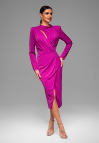 Fuchsia jurk met lange mouwen, een wikkelontwerp en asymmetrische uitsparingen, met een split tot boven de dij en gedrapeerde details. Zilveren bandjeschoenen.