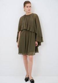 Olijfgroene chiffon jurk met gelaagde capesleeven, ronde hals en een vloeiende silhouet, aangevuld met zwarte puntige ballerina's en gouden oorbellen.