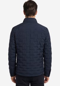 Mann trägt eine marineblaue gesteppte Jacke, steht mit dem Rücken zur Kamera und zeigt den Rücken sowie lange Ärmel mit geknöpften Manschetten.