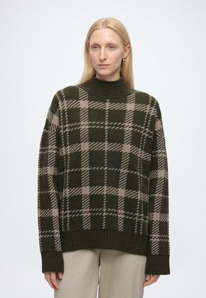 Femme blonde portant un pull à carreaux vert foncé et beige à col montant et un pantalon beige, debout devant un fond uni.