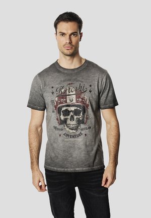 Camiseta gris de manga corta con un gráfico de una calavera con casco rojo, con texto y motivos de rayos. Corte casual, textura suave.