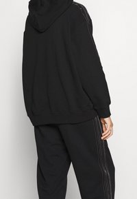 Černá oversize mikina s kapucí, zdobená vertikálními stehujícími detaily na rukávech. Kombinována s volnými černými tepláky, také s detaily stehů.
