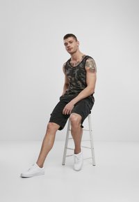 Un maiou negru cu model camuflaj, purtat împreună cu pantaloni scurți cargo, așezat pe un scaun. Adidași albi completează ținuta. Design simplu și aspect casual.