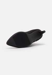Zapato de tacón alto de ante negro con puntera afilada y suela de goma texturizada. Presenta un diseño sin cordones y acentos minimalistas.