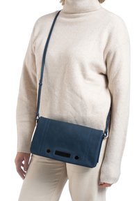 Blauwe leren schoudertas met contrasterende stiksels, rechthoekige vorm, gaasaccent en verstelbare schouderband, gedragen met een licht sweater.