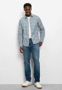 Weißes und blau gemustertes Hemd mit langen Ärmeln, getragen über einem hellen Hemd, kombiniert mit hellblauen Jeans und weißen Turnschuhen, steht vor einem schlichten Hintergrund.