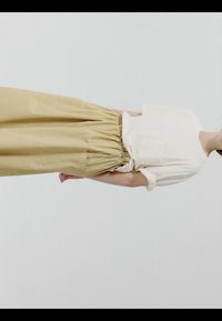 Personne portant une chemise blanche à manches courtes rentrée dans un pantalon beige à taille haute et jambes larges, sur un fond clair uni.