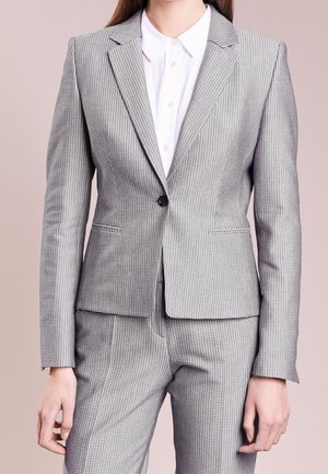 Kvinde iført en grå jakkesæt med hundetandsmønster, med en enkel-knaps blazer og matchende bukser, kombineret med en hvid skjorte med knapper.
