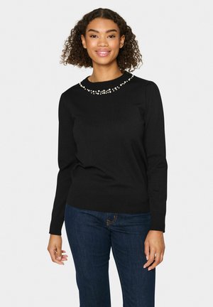 Maglione nero con maniche lunghe e scollatura texturizzata accentuata da perline bianche. La vestibilità è aderente con orlo a coste. Indossato con jeans scuri.