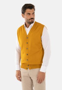 Gilet lavorato a maglia giallo con scollatura a V e cinque bottoni scuri, indossato sopra una camicia bianca e abbinato a pantaloni beige. Tessuto liscio.