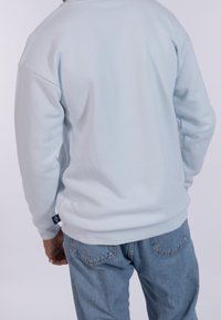 Felpa leggera di colore blu chiaro, di peso medio, con collo rotondo e maniche lunghe. La vista posteriore mostra una trama liscia e un dettaglio con etichetta laterale. Indossata con jeans blu.