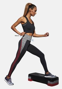 Czarne legginsy sportowe z szarym pasem i bordowymi akcentami, noszone z czarnym stanikiem sportowym. Tło jest jednolicie białe.