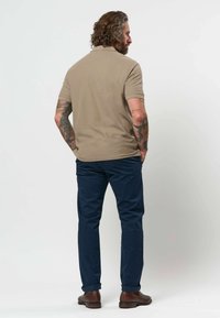 Homme avec des tatouages portant un polo beige à manches courtes, un pantalon bleu marine et des chaussures marron, debout de dos, les mains dans les poches sur fond blanc.