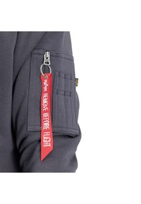 Šedá mikina se zipovou kapsou a červeným štítkem "ODSTRANĚT PŘED LETEM". Vyznačuje se strukturovanou tkaninou a dalšími bočními prošívacími detaily.